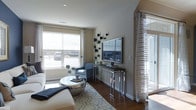 This single-floor space showcases a thoughtfully designed living area that blends contemporary style with comfortable functionality. The living room features rich hardwood flooring anchored by a patterned area rug in neutral tones with blue accents, complementing the overall color palette of the space. Large windows fitted with roller shades and sheer, textured curtain panels allow generous natural light to fill the room, creating a bright and welcoming atmosphere. Sheer floor-length drapery near the entryway adds an elegant, airy touch.

The room is furnished with a clean-lined sofa and a coordinating blue patterned accent chair, arranged around a distinctive round metallic coffee table that serves as a modern focal point. A sleek, chrome-framed media console holds a flat-screen television mounted against the wall, accented by an arrangement of decorative wall art in deep blue and charcoal tones. Recessed lighting in the ceiling provides additional illumination. The neutral gray walls and carefully curated accessories give the space a polished, cohesive aesthetic suited for both relaxing and entertaining.