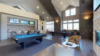 This single-floor clubhouse is a well-appointed social and recreational hub designed with both style and function in mind. The space features soaring vaulted ceilings that create an open, airy atmosphere, complemented by an abundance of natural light streaming through a striking arrangement of large upper-level windows. The interior design blends contemporary finishes with warm, inviting textures throughout.

At the heart of the space is a spacious game room area, anchored by a full-size billiards table and a shuffleboard table, offering ample room for recreation and entertainment. A dedicated bar nook, framed by natural stone accent walls and a mosaic tile backsplash, provides a stylish setting complete with bar-height seating and wall-mounted flat-screen televisions — ideal for gathering during events or game days.

The lounge area features modern curved seating, including plush accent chairs and a sleek side table, creating an intimate space for conversation and relaxation. Rich wood-look flooring runs throughout, tying together the various zones of the clubhouse seamlessly. Recessed lighting adds warmth and dimension to the vaulted ceiling, enhancing the overall ambiance. This clubhouse delivers a sophisticated yet comfortable environment suited for a wide range of social activities.