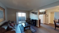 This single-floor unit offers an open and functional layout that seamlessly connects the living area to the kitchen. The living room is anchored by hardwood-style flooring and features a sliding glass door that fills the space with natural light and provides access to a private balcony overlooking a covered parking area and surrounding residential buildings. A textured ceiling adds depth overhead, while neutral-toned walls create a versatile backdrop throughout. The living area comfortably accommodates seating furniture and a coffee table, with ample room to move freely. A partial wall with an archway separates the living space from the kitchen, which is outfitted with dark cabinetry, a white refrigerator, and a glimpse of counter workspace. The kitchen benefits from its own overhead lighting and connects naturally to the main living space, making it well-suited for everyday use. The balcony, accessible through the sliding door, offers an outdoor retreat with views of the surrounding complex and open sky. Overall, the unit presents a straightforward, well-lit layout with a practical flow between its key living and cooking areas.