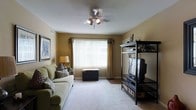 This single-floor space offers a welcoming and functional living environment with a warm, homey ambiance. The living room serves as the heart of the space, featuring plush wall-to-wall carpeting that adds comfort underfoot and contributes to the room's cozy atmosphere. Warm golden-beige walls create an inviting backdrop, complemented by neutral-toned décor throughout.

A large window dressed with vertical blinds and decorative curtain panels floods the room with natural light, making the space feel open and airy. A ceiling fan with an integrated light fixture is centrally positioned overhead, providing both practical ventilation and ambient lighting.

The living room is well-appointed with comfortable seating, including a green upholstered sofa accented with decorative pillows. An open black shelving unit houses a television and provides additional storage, while framed wall art adds a personal touch to the space. A decorative storage trunk beneath the window offers both style and functionality.

The layout is practical and well-organized, maximizing the use of available floor space. Overall, this single-floor residence presents a comfortable, move-in-ready living environment with classic, understated décor and a bright, welcoming atmosphere.