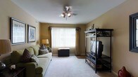 This single-floor space offers a welcoming and functional living environment with a warm, homey ambiance. The living room serves as the heart of the space, featuring plush wall-to-wall carpeting that adds comfort underfoot and contributes to the room's cozy atmosphere. Warm golden-beige walls create an inviting backdrop, complemented by neutral-toned décor throughout.

A large window dressed with vertical blinds and decorative curtain panels floods the room with natural light, making the space feel open and airy. A ceiling fan with an integrated light fixture is centrally positioned overhead, providing both practical ventilation and ambient lighting.

The living room is well-appointed with comfortable seating, including a green upholstered sofa accented with decorative pillows. An open black shelving unit houses a television and provides additional storage, while framed wall art adds a personal touch to the space. A decorative storage trunk beneath the window offers both style and functionality.

The layout is practical and well-organized, maximizing the use of available floor space. Overall, this single-floor residence presents a comfortable, move-in-ready living environment with classic, understated décor and a bright, welcoming atmosphere.