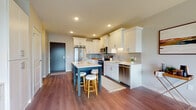 This single-floor residence offers a thoughtfully designed open-concept layout with warm hardwood-style flooring flowing throughout the main living areas. The kitchen is a standout feature, showcasing crisp white shaker cabinetry with crown molding detail, a grey subway tile backsplash, light quartz countertops, and a full suite of stainless steel appliances including a French door refrigerator, range, microwave, and dishwasher. A two-toned island with a navy base and white countertop provides seating for two and serves as the centerpiece of the space, ideal for casual dining or entertaining. Recessed lighting illuminates the kitchen and living areas, contributing to a bright, inviting atmosphere. The living room features a neutral color palette with large windows and a sliding glass door that opens to a private balcony, allowing for generous natural light and outdoor views. The bedroom is carpeted and well-lit by a large window, offering a serene retreat with ample space. The bathroom connects directly to the bedroom, featuring clean, simple finishes with hardwood-style flooring and a functional layout. Overall, the space presents a modern, cohesive aesthetic suited for comfortable everyday living.