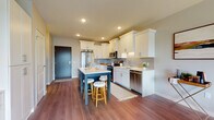 This single-floor residence offers a thoughtfully designed open-concept layout with warm hardwood-style flooring flowing throughout the main living areas. The kitchen is a standout feature, showcasing crisp white shaker cabinetry with crown molding detail, a grey subway tile backsplash, light quartz countertops, and a full suite of stainless steel appliances including a French door refrigerator, range, microwave, and dishwasher. A two-toned island with a navy base and white countertop provides seating for two and serves as the centerpiece of the space, ideal for casual dining or entertaining. Recessed lighting illuminates the kitchen and living areas, contributing to a bright, inviting atmosphere. The living room features a neutral color palette with large windows and a sliding glass door that opens to a private balcony, allowing for generous natural light and outdoor views. The bedroom is carpeted and well-lit by a large window, offering a serene retreat with ample space. The bathroom connects directly to the bedroom, featuring clean, simple finishes with hardwood-style flooring and a functional layout. Overall, the space presents a modern, cohesive aesthetic suited for comfortable everyday living.