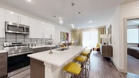 Located at 4500 Kittredge St in Denver, Colorado, this well-appointed one-bedroom, one-bathroom residence offers a thoughtfully designed single-floor layout. The open-concept kitchen and living area serves as the heart of the home, featuring sleek white upper cabinetry, dark wood lower cabinets, a tiled backsplash, and a spacious waterfall-edge island with bar seating — ideal for both cooking and casual dining. Stainless steel appliances, including a refrigerator, range, microwave, and dishwasher, complete the modern kitchen. Wood-look flooring runs throughout the main living areas, while recessed and pendant lighting add warmth and depth. The living room offers a comfortable, light-filled retreat with large windows dressed in floor-to-ceiling drapes. The bedroom provides a cozy, carpeted sanctuary with ample natural light, a sliding barn door leading to the closet, and generous window treatments for privacy. The bathroom is finished with large-format tile flooring, a walk-in glass shower, a floating dark wood vanity with quartz countertop, open shelving, and a backlit mirror — delivering a clean, spa-like aesthetic. This residence combines contemporary finishes with a functional layout, offering comfortable urban living in Denver.