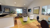 This single-floor space is defined by its striking vaulted ceilings, which lend an airy, expansive quality to the open-concept layout. Warm hardwood flooring flows throughout, unifying the living, dining, and kitchen areas into a cohesive and welcoming environment.

The living area is thoughtfully arranged with a grey sofa accented by patterned throw pillows, two contemporary white barrel chairs, and a glass coffee table set atop a vibrant chartreuse area rug. A wall-mounted flat-screen television and framed artwork contribute to the polished aesthetic, while a wall-mounted mini-split system ensures year-round climate comfort. Natural light enters through slatted windows overlooking greenery.

The dining area features a modern glass-top table with sleek black chairs, positioned near a glass-paneled entry door and wide windows that frame outdoor foliage, creating a bright and inviting atmosphere.

The kitchen is equipped with rich dark cabinetry, granite countertops, a stainless steel side-by-side refrigerator with ice and water dispenser, a built-in electric cooktop, and an over-the-range microwave. A pass-through counter connects the kitchen to the adjacent living space, enhancing the open flow. A compact bistro-style table with bar stools provides an additional casual dining option nearby.