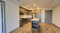 This contemporary single-floor space showcases a thoughtfully designed open-concept layout that maximizes both functionality and style. The kitchen serves as the heart of the space, featuring flat-panel wood-toned cabinetry that extends to the ceiling, offering generous storage throughout. A full suite of stainless steel appliances — including a gas range, over-the-range microwave, and refrigerator — provides a modern, professional-grade cooking experience. The crisp white countertops contrast beautifully against the warm cabinetry, while a freestanding central island with open lower shelving adds additional prep and storage space, doubling as a casual dining or entertaining surface. Light hardwood flooring runs throughout, contributing to a warm and inviting ambiance that complements the clean white walls. Recessed lighting illuminates the space evenly, enhancing its bright, airy feel. The open layout creates a seamless flow between the kitchen and adjacent living areas, making the space feel expansive and well-connected. The overall design strikes a balance between contemporary aesthetics and practical living, with quality finishes and a neutral palette that offers versatile decorating possibilities.