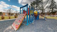 Located at 5750 Roche Dr in Columbus, Ohio, this outdoor playground serves as a recreational amenity within a residential community. The play area is surfaced with gravel and centers around a vibrant, multi-component play structure framed in bold blue, red, and yellow. The structure offers a variety of engaging features for children, including a rock climbing wall with colorful hand grips, a tire climbing panel, ladder rungs, a yellow enclosed tunnel slide, wooden ramps, and multiple elevated platforms connected by various access points. Alongside the main structure, a swing set with green belt swings supported by red metal posts provides additional play options. A paved walkway borders the gravel play surface, leading toward a grassy open area where two gazebos are visible in the background, suggesting additional outdoor gathering space for the community. Mature trees are scattered throughout the surrounding grounds, providing a natural backdrop and seasonal shade. The playground is set against the backdrop of multi-story brick residential buildings, reinforcing its role as a community-facing amenity. The overall layout is open and accessible, offering a well-rounded outdoor play environment.