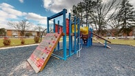 Located at 5750 Roche Dr in Columbus, Ohio, this outdoor playground serves as a recreational amenity within a residential community. The play area is surfaced with gravel and centers around a vibrant, multi-component play structure framed in bold blue, red, and yellow. The structure offers a variety of engaging features for children, including a rock climbing wall with colorful hand grips, a tire climbing panel, ladder rungs, a yellow enclosed tunnel slide, wooden ramps, and multiple elevated platforms connected by various access points. Alongside the main structure, a swing set with green belt swings supported by red metal posts provides additional play options. A paved walkway borders the gravel play surface, leading toward a grassy open area where two gazebos are visible in the background, suggesting additional outdoor gathering space for the community. Mature trees are scattered throughout the surrounding grounds, providing a natural backdrop and seasonal shade. The playground is set against the backdrop of multi-story brick residential buildings, reinforcing its role as a community-facing amenity. The overall layout is open and accessible, offering a well-rounded outdoor play environment.