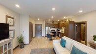 This single-floor unit offers a thoughtfully designed open-concept layout that seamlessly connects the living, dining, and kitchen areas. Light wood-style flooring runs throughout the main living spaces, creating a warm, cohesive aesthetic complemented by neutral wall tones and recessed lighting overhead.

The kitchen is a standout feature, equipped with rich dark-brown shaker-style cabinetry extending to the ceiling, granite-look countertops, a white subway tile backsplash, and a full suite of stainless steel appliances including a refrigerator, electric range, microwave, and dishwasher. A compact dining area with a round table sits adjacent to the kitchen, ideal for everyday meals.

The living room is bright and welcoming, featuring large windows that allow generous natural light and views of outdoor greenery. A private entry door is accessible directly from this space.

The bedroom is carpeted for comfort, features a ceiling fan, a window with outdoor views, and connects to a full bathroom equipped with a granite-topped double vanity, dark wood cabinetry, and a shower. A second bathroom mirrors the same design quality, featuring a single vanity with granite countertop and a walk-in shower, providing added convenience.