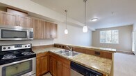 This single-floor unit offers a functional and efficiently organized layout comprising a living room, kitchen, bedroom, hallway, and closet. The kitchen serves as a central highlight, featuring warm medium-toned wood cabinetry, granite-style countertops with tile accents, a stainless steel refrigerator, electric range, over-the-range microwave, double-basin sink, and a dishwasher. Light wood-look flooring runs throughout the kitchen, while pendant lighting suspended above the peninsula adds a refined touch. The open sightline from the kitchen toward the living area enhances the sense of space.

The living room is carpeted in neutral tones with light-colored walls, providing a clean and versatile canvas. A door with sidelights allows natural light to filter in and provides outdoor access. The bedroom is a comfortable, well-proportioned space with carpet flooring, neutral walls, and a window offering outdoor views. The walk-in closet off the bedroom includes built-in shelving units and dual hanging rods, providing substantial and organized storage. The hallway connects the rooms efficiently, contributing to the overall practical flow of the space.