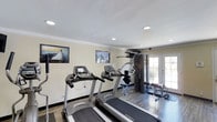 This single-floor fitness center is designed to support a well-rounded workout routine. The space is outfitted with multiple treadmills, including Spirit-branded models, positioned along the wall for convenient cardio training. A multi-station cable and weight machine anchors the strength-training area, complemented by a rack of free weights, providing users with a comprehensive range of exercise options.

The interior finishes lend the space a polished, well-maintained appearance. Crown molding lines the ceiling, which features recessed lighting that illuminates the room evenly. The flooring consists of wood-look planks that add warmth and durability underfoot. The walls are painted in a neutral tone and are accented by motivational fitness posters, contributing to an energizing atmosphere.

Natural light enters through a set of French doors at the far end of the room, offering a connection to the outdoors and enhancing the overall brightness of the space. The layout is open and functional, allowing for ease of movement between equipment. Overall, this fitness center combines practical amenities with refined architectural details to create an inviting and effective exercise environment.