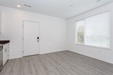 This single-floor unit presents a clean, modern aesthetic defined by a neutral color palette, light hardwood-style flooring throughout, and crisp white walls. The open-concept layout allows the kitchen and living areas to flow seamlessly together, creating a spacious and functional environment.

The kitchen is well-appointed with white shaker-style cabinetry reaching toward the ceiling, offering ample storage alongside open display shelving. Dark countertops provide a striking contrast to the light cabinetry, and the suite of stainless steel appliances — including a refrigerator, electric range, built-in microwave, and dishwasher — adds a polished, contemporary feel. A single-basin stainless steel sink with a modern faucet completes the setup.

The living area benefits from natural light streaming through window blinds, maintaining privacy while brightening the space. The entryway features a keypad-secured front door alongside an additional window, enhancing both security and light. Bedroom areas are calm and understated, with neutral taupe walls and continued hardwood-style flooring.

The bathroom is finished in an all-white scheme, featuring a vanity with white cabinetry, a toilet, and a tub-shower combination with a curved rod. Recessed lighting throughout the unit contributes to an overall bright and welcoming atmosphere.