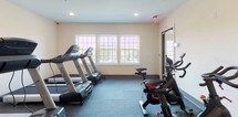 This fitness center is a thoughtfully designed, single-floor space built to support a comprehensive workout experience. The layout is open and functional, allowing for comfortable movement between distinct exercise zones. Recessed ceiling lighting illuminates the space evenly throughout, while a row of large windows along one wall invites natural light and offers views of the surrounding greenery, creating an energizing atmosphere.

The cardio zone is well-appointed with multiple Spirit treadmills and an elliptical trainer arranged in a neat row, complemented by BodyCraft cycling bikes positioned nearby. A full multi-station strength training machine occupies its own dedicated area, providing a wide range of resistance and weight training options. The free weights section features a tiered dumbbell rack stocked with a full set of weights, alongside an adjustable workout bench for targeted strength exercises.

The flooring throughout is durable rubber gym matting in a dark gray tone, offering both safety and cushioning underfoot. The neutral, light-toned walls and clean white paneled doors contribute to a bright and professional environment. An emergency exit sign is visible, reflecting attention to safety standards. Overall, this fitness center is a practical and well-maintained space designed to meet a variety of fitness needs.