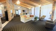 This single-story space offers a welcoming and open atmosphere defined by its striking architectural character. The vaulted ceiling, finished with warm wood paneling and supported by exposed beams, draws the eye upward and contributes to an expansive sense of height. Multiple ceiling fans are evenly distributed throughout, ensuring comfortable airflow across the generous floor area.

The exposed brick walls add texture and a rustic warmth to the interior, complementing the neutral-toned carpeting that runs throughout. Large vertical windows and sliding glass doors allow natural light to pour into the space, creating a bright and airy environment while offering views to an outdoor area.

The lounge is furnished with cream-colored sofas accented by colorful throw pillows, a glass-top coffee table, and upholstered armchairs arranged to encourage conversation and relaxation. A wall-mounted flat-screen television anchors one seating area, while a floor lamp and decorative greenery add finishing touches. The overall ambiance is casual yet polished, making this a comfortable and functional gathering space suitable for socializing or unwinding.