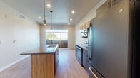 This well-appointed single-floor unit offers a thoughtfully designed layout with contemporary finishes throughout. The open-concept kitchen serves as the centerpiece of the space, featuring a large center island with a built-in sink, sleek gray quartz countertops, and light wood cabinetry that provides generous storage. A full suite of stainless steel appliances — including a refrigerator, electric range, built-in microwave, and dishwasher — completes the kitchen. Pendant lighting above the island and recessed ceiling lights add both style and functionality. The kitchen flows naturally toward a sliding glass door that opens to an outdoor area, allowing natural light to fill the space. Warm hardwood-style flooring runs throughout the main living areas, creating a cohesive and inviting atmosphere. The bedroom is bright and airy, benefiting from a large window framing picturesque views of the surrounding landscape, including distant mountains and greenery. The bathroom is clean and functional, featuring a full soaking tub with a tiled surround, a stylish teal vanity with a quartz countertop, and recessed lighting. The neutral color palette and modern design elements throughout give the space a fresh, move-in-ready feel.