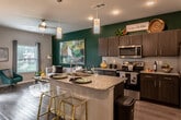 This well-appointed single-floor residence showcases a thoughtfully designed open-concept layout where the kitchen flows seamlessly into the living area, creating a bright and cohesive space. The kitchen is a standout feature, boasting dark espresso cabinetry, granite countertops, a central island with an undermount sink and pendant lighting above, and a full suite of stainless steel appliances including a refrigerator, range, and microwave. A bold emerald green accent wall anchors the kitchen and living area, adding a vibrant, contemporary energy throughout the space. Wood-style flooring runs through the main living areas, while a ceiling fan and large windows allow natural light to fill the room. The living area is open and inviting, with outdoor views accessible through sliding glass doors. The primary bedroom features a warm yellow accent wall, plush carpeting, and a ceiling fan, creating a cozy and restful retreat. A second bedroom offers neutral tones, natural light from a window, and comfortable carpeted flooring. The bathroom is finished with clean white subway tile surrounding a soaking tub and shower combination, complemented by dark cabinetry for a modern contrast. Overall, the space strikes a balance between bold design choices and functional, comfortable living.