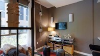 This single-floor space showcases a carefully curated, eclectic interior design that blends urban sophistication with warm, inviting elements. The living area features rich, earthy taupe-toned walls that create a cozy yet stylish atmosphere, complemented by dark hardwood flooring that runs throughout. Natural light streams in through a large window dressed with elegant white curtains, offering views of the surrounding cityscape and adding a bright, airy quality to the space. Wall-mounted sconce lighting provides a soft, ambient glow, enhancing the room's intimate character. A wall-mounted flat-screen television serves as a focal point, positioned above a distinctive mirrored and metal console table adorned with woven rattan baskets for practical storage. The decor includes charming macramé hanging planters with lush greenery, metallic sculptural accents, and eclectic stools that contribute to the space's layered, artistic personality. A vibrant area rug anchors the seating arrangement, adding a pop of color against the darker tones of the room. Every detail reflects intentional design choices, resulting in a space that feels both functional and visually dynamic, with a personality that blends industrial, bohemian, and contemporary influences seamlessly.