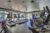 Located at 100 Perimeter Trace E in Dunwoody, Georgia, this well-appointed fitness center offers a comprehensive workout environment designed for both comfort and functionality. The space is defined by its elegant arched windows trimmed in dark wood, which flood the interior with abundant natural light and provide views of the surrounding outdoor greenery. The rubber-floored workout area is outfitted with a full suite of Matrix-brand cardio equipment, including multiple treadmills and elliptical cross-trainers, each equipped with individual display screens. A row of elliptical machines is positioned along a mirrored wall, which visually expands the space and allows users to monitor their form during exercise. Ceiling fans are installed throughout, ensuring consistent airflow and comfort during workouts. Additional fitness accessories are readily available, including kettlebells of varying weights, stability balls, medicine balls, and a yoga mat, supporting a diverse range of training routines. Recessed lighting complements the natural light from the windows, keeping the space well-illuminated at all times. A fitness information board is mounted near the entrance, providing guidance for users. The overall ambiance is clean, polished, and inviting, combining practical fitness amenities with a refined, upscale aesthetic suited to the Dunwoody community.