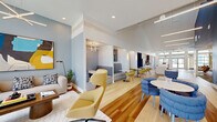 Located at 98 Northwood Blvd in Pittsboro, NC, this single-floor community clubhouse offers a thoughtfully designed gathering space that blends contemporary style with functional amenities. The open-plan layout is anchored by soaring double-height ceilings and warm hardwood flooring that flows throughout, creating a bright and welcoming atmosphere.

The main lounge area serves as the heart of the space, featuring a prominent branded wood-slat accent wall, bold navy blue accents, and an eclectic mix of modern furnishings including sculptural lounge chairs, soft seating groupings, and a large central area rug. Floor-to-ceiling windows and double glass entry doors flood the interior with natural light while offering views of the surrounding community.

A fully equipped kitchen includes stainless steel appliances, warm wood cabinetry, a large white quartz island with bar seating, and an adjacent built-in banquette dining nook illuminated by a geometric chandelier — ideal for casual gatherings or community events.

The social lounge transitions seamlessly into a variety of flexible seating arrangements, including café-style tables and upholstered booth seating. A dedicated conference room provides a more private setting, outfitted with a long communal table, ergonomic seating, wall-mounted display screen, and distinctive hexagonal acoustic panels that add both function and visual interest. Recessed and pendant lighting throughout ensures a polished, well-lit environment in every zone.