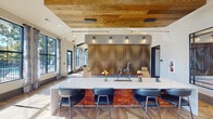 Located at 5715 Glenwood Ave in Raleigh, NC, this single-floor clubhouse showcases a sophisticated blend of rustic warmth and contemporary design. The space is anchored by striking herringbone and geometric-patterned hardwood flooring that flows throughout, complemented by exposed wood ceiling beams and reclaimed wood wall accents that add rich texture and character.

The main lounge area features soaring vaulted ceilings, oversized floor-to-ceiling windows that flood the interior with abundant natural light, and plush sectional seating arranged around a large-scale landscape mural. A shuffleboard table and oversized interactive game board add recreational appeal. A welcoming gathering nook is appointed with tufted leather wingback chairs, a rustic chandelier with Edison bulb styling, a wall-mounted display, and a sleek linear fireplace — all framed by panoramic outdoor views.

A well-equipped kitchen bar area offers a butcher block island with bar seating, cabinetry with reclaimed wood detailing, a beverage refrigerator, and a coffee station. The main reception area features a long white quartz bar with modern stools backed by a dramatic fluted wood accent wall. Private workspaces include built-in desks positioned beneath large windows overlooking lush greenery, providing a focused and naturally lit environment. The entry foyer showcases intricate inlaid hardwood flooring and generous window frontage, creating a bright and welcoming arrival experience.