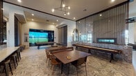 This single-story lounge and game room in Cypress, TX, is a thoughtfully designed social space that blends upscale aesthetics with entertainment-focused amenities. The layout flows seamlessly between distinct activity zones, unified by light wood-style flooring and eye-catching patterned tile in the central gathering area.

The game room section is well-appointed, featuring a full-size billiards table with hairpin-style legs, a shuffleboard table positioned along a decoratively tiled half-wall, a ping-pong table, and a glass-enclosed golf simulator bay with a projector and putting surface. Tall, upholstered wall panels add acoustic warmth and visual texture near the game area.

The lounge area offers a refined atmosphere, anchored by a tufted velvet sofa and complemented by a striking floor-to-ceiling marble-look accent wall. A built-in horizontal aquarium is integrated directly into the wall, serving as a unique focal point. Ornate gold-framed mirrors and vintage-style lantern sconces add an elegant touch. Marble-top pedestal tables provide intimate seating options nearby.

A bar counter with stools runs along one wall, and a wall-mounted television is positioned for communal viewing. Sculptural pendant lighting throughout ties the space together, while large windows invite natural light and outdoor views into the lounge area.