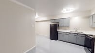 This single-floor unit offers a clean, functional layout designed for practical everyday living. The kitchen serves as a central feature of the space, equipped with gray shaker-style cabinetry spanning both upper and lower sections, a white countertop, a stainless steel sink, and a full suite of black appliances including a refrigerator, range, and dishwasher. Flush-mounted ceiling lights provide bright, even illumination throughout the kitchen area, which flows openly into the adjacent living space. Consistent large-format white tile flooring runs throughout, lending a cohesive and easy-to-maintain aesthetic across the main areas. The bedroom is a comfortably proportioned room featuring the same tile flooring, neutral beige walls, a ceiling fan with integrated lighting, and a window dressed with white horizontal blinds that allows natural light to filter in. A secondary windowed room offers additional flexible space, also finished with light-toned walls and tile flooring. The bathroom is straightforward and well-appointed, featuring a tiled tub and shower combination with a curved curtain rod, a toilet, and a sink. Crown molding accents portions of the interior, adding a subtle architectural detail. Overall, the space presents a tidy, move-in ready environment with a neutral palette throughout.