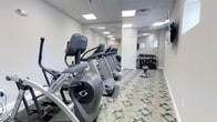 This fitness center offers a clean, functional layout designed to accommodate a range of exercise routines. The space is outfitted with a lineup of Cybex Arc Trainer elliptical machines and treadmills arranged in a row along one wall, providing ample cardio equipment for users. A full-length mirror spans one wall, enhancing the sense of space while allowing users to monitor their form during workouts.

The free weight area is situated toward the back of the room, featuring a well-stocked dumbbell rack with an accompanying weight bench, suitable for strength training exercises. The flooring consists of a patterned, cushioned surface that provides both comfort and durability underfoot.

Illumination is provided by recessed panel lighting set into a drop ceiling, creating a bright and evenly lit environment throughout. The walls are finished in a neutral light grey, contributing to the room's clean and modern aesthetic. A small window near the free weight area allows for a modest amount of natural light. The overall ambiance is practical and inviting, making it a capable facility for daily fitness needs.