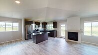 This single-floor home offers a thoughtfully designed open-concept layout finished with light wood-tone flooring that extends seamlessly throughout. The kitchen serves as the centerpiece of the main living area, showcasing rich dark espresso cabinetry paired with polished dark granite countertops. A substantial center island with a built-in sink provides additional workspace and casual gathering space. Stainless steel appliances, including a French door refrigerator, electric range, and over-the-range microwave, complete the kitchen's functional setup. Multiple windows along the kitchen wall frame open rural views, filling the space with natural light. Adjacent to the kitchen, the living area features a sleek contemporary fireplace and direct access to an exterior door, with an expansive open floor plan providing generous living space.

The bedrooms are well-proportioned, each featuring vaulted or elevated ceilings, ceiling fans, baseboard heating, and large windows overlooking surrounding green fields. Sliding panel closets provide ample storage. The bathrooms are finished with matching dark granite vanity countertops, undermount sinks, and clean white tub-shower combinations, maintaining a consistent modern aesthetic throughout the home.