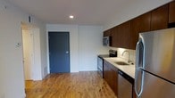 This single-floor space showcases a clean, modern design anchored by warm hardwood flooring that runs throughout the open layout. The kitchen is a standout feature, outfitted with rich, dark wood cabinetry that extends from counter height to the ceiling, maximizing storage capacity. Stainless steel appliances — including an electric range, built-in microwave, and dishwasher — complement the light-toned countertops, which also house a single-basin sink with a contemporary gooseneck faucet. The white subway tile backsplash adds a crisp, clean contrast to the darker cabinetry. The open area adjacent to the kitchen provides a versatile living space, kept bright and neutral with white walls and recessed lighting overhead. A bold slate-blue entry door adds a subtle design accent and provides a clear sense of entry and separation. The overall aesthetic balances functional, well-organized kitchen space with an open, unfussy living area, creating a practical and visually cohesive environment throughout.