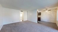 This single-floor residence offers a straightforward and functional layout designed for comfortable everyday living. The main living area is generously proportioned, featuring wall-to-wall plush carpeting that adds warmth underfoot and a soft, neutral palette throughout. The textured ceiling and light-toned walls create a clean, understated aesthetic that allows for versatile interior styling.

A centrally positioned opening connects the living space to the kitchen, which is equipped with dark wood cabinetry providing ample storage. The kitchen transitions to a hard-surface floor, offering a practical contrast to the carpeted living zone, and includes counter space suited for daily meal preparation.

A hallway visible from the living area leads to additional rooms, with natural light filtering through from further within the home, suggesting well-placed windows that brighten the interior. A ceiling fan is visible in an adjoining area, contributing to airflow and comfort throughout the space.

Overall, the residence presents a bright, open, and move-in ready environment with a simple, cohesive design. The neutral finishes and practical layout make it an adaptable space well-suited to a variety of furnishing styles and living arrangements.