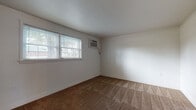 This single-floor space offers a practical and comfortable layout designed for everyday living. The floor plan, as revealed in the dollhouse view, includes multiple rooms connected by a straightforward arrangement that maximizes usability.

The bedrooms are generously proportioned, each finished with warm brown carpeting and crisp white walls. Multiple windows fitted with horizontal blinds allow ample natural light to fill each room, creating a bright and airy atmosphere. One of the rooms also features a wall-mounted air conditioning unit, providing direct climate control. Clean white baseboards frame each carpeted space neatly throughout.

The kitchen is well-equipped and functional, featuring a full run of warm oak cabinetry extending from floor to ceiling, offering substantial storage. Dark-toned countertops provide a grounded contrast to the light cabinetry, while white appliances — including an electric range and refrigerator — are included. Stone-look tile flooring adds durability to this high-traffic area. A window above the range area brings in natural light, keeping the kitchen feeling open.

The bathroom includes a full-size soaking tub with a tiled surround and built-in corner shelving, providing practical storage within the shower/tub enclosure. Overall, the space presents a clean, move-in-ready environment with consistent finishes throughout.