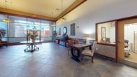 Located at 130 Rollins Ave in Rockville, Maryland, this single-floor commercial lobby presents a polished and welcoming environment designed to impress. The space is anchored by large-format slate tile flooring laid in a diagonal pattern, lending a sense of depth and sophistication throughout. Soaring ceilings with crown molding details and warm wood-toned trim accents contribute to the grand, refined atmosphere.

The lobby is thoughtfully curated with decorative artwork, including richly colored framed prints displayed along the walls, complemented by striking bronze sculptures that serve as focal points. Seating areas feature elegantly upholstered armchairs with floral and leaf motifs, arranged around small side tables to create comfortable gathering spots. Decorative vases and potted plants add organic warmth to the interior.

A generous lounge area near the floor-to-ceiling glass storefront allows abundant natural light to flood the space, offering views of the surrounding streetscape. Additional seating with dining-height tables and cushioned chairs provides flexible use options. Pendant and flush-mount light fixtures with decorative shades enhance the ambient lighting throughout.

The elevator bank, framed by wainscoting and coordinating artwork, is accessible from the main lobby area, completing a well-appointed and functional space suitable for a professional office or mixed-use building.