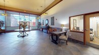 Located at 130 Rollins Ave in Rockville, Maryland, this single-floor commercial lobby presents a polished and welcoming environment designed to impress. The space is anchored by large-format slate tile flooring laid in a diagonal pattern, lending a sense of depth and sophistication throughout. Soaring ceilings with crown molding details and warm wood-toned trim accents contribute to the grand, refined atmosphere.

The lobby is thoughtfully curated with decorative artwork, including richly colored framed prints displayed along the walls, complemented by striking bronze sculptures that serve as focal points. Seating areas feature elegantly upholstered armchairs with floral and leaf motifs, arranged around small side tables to create comfortable gathering spots. Decorative vases and potted plants add organic warmth to the interior.

A generous lounge area near the floor-to-ceiling glass storefront allows abundant natural light to flood the space, offering views of the surrounding streetscape. Additional seating with dining-height tables and cushioned chairs provides flexible use options. Pendant and flush-mount light fixtures with decorative shades enhance the ambient lighting throughout.

The elevator bank, framed by wainscoting and coordinating artwork, is accessible from the main lobby area, completing a well-appointed and functional space suitable for a professional office or mixed-use building.