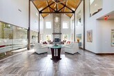Located in Layton, Utah, this well-appointed two-floor facility offers an impressive range of amenities designed for recreation, fitness, and social gathering. The main floor is the larger of the two levels and serves as the heart of the space. A dramatic living/lounge area features soaring ceilings, exposed wood beam accents, and a floor-to-ceiling stacked stone fireplace that anchors the room with warmth and character. Large windows flood the space with natural light, while glass panel walls provide visibility between zones. A dedicated home theater room is outfitted with tiered rows of leather reclining seats, a ceiling-mounted projector, and movie poster décor for an immersive cinematic experience. The kitchenette features dark wood cabinetry, stainless steel appliances, stone tile flooring, and a window overlooking the outdoors. The upper floor houses a well-equipped fitness center with treadmills, cable machines, free weights, and wall mirrors beneath striking coffered wood-beam ceilings and abundant natural light. A spacious game room on this level offers room for a full-size pool table and ping pong, complemented by wide windows with outdoor views. A meeting or lounge area with exposed beam vaulted ceilings, a round table, and interior glass windows rounds out the upper level's versatile offerings.