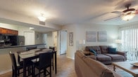 This single-floor residence offers a thoughtfully designed layout that balances comfort and functionality throughout. The open-concept living and dining area serves as the heart of the home, where wood-look flooring, a ceiling fan, and a tile-surround fireplace create a warm, inviting atmosphere. A sliding glass door leads to an outdoor balcony, allowing natural light to fill the space. The dining area accommodates a round table with seating, positioned conveniently adjacent to the kitchen. The kitchen is well-equipped with rich cherry-toned cabinetry, dark granite-style countertops, black appliances including a refrigerator, range, and microwave, as well as a double-basin sink and dishwasher. The primary bedroom features a queen-sized bed, matching nightstands, and direct access to an en-suite bathroom complete with a wide vanity, dark countertops, and ample storage. A second bedroom offers two twin beds, a natural light-filled window, and closet access. The second full bathroom mirrors the first in style, featuring a similarly appointed vanity and shower. Wood-look flooring runs consistently throughout, tying the residence together with a cohesive, modern aesthetic.