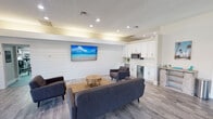 This single-floor community clubhouse offers a thoughtfully designed space that blends contemporary style with a relaxed coastal aesthetic. The main entrance welcomes visitors into a bright, open reception area featuring large front-facing windows that flood the space with natural light and offer views of the surrounding exterior. A sleek, wood-toned reception desk anchors the area, complemented by light hardwood-style flooring that runs throughout the entire facility.

The central lounge is a standout feature, showcasing a shiplap accent wall in crisp white that serves as the backdrop for a wall-mounted flat-screen television. Comfortable mid-century modern seating, including a large sofa and upholstered armchairs, is arranged around a natural wood coffee table atop a woven area rug, creating an inviting gathering space. Recessed lighting and an indirect cove light detail along the ceiling add warmth and depth to the room.

Adjacent to the lounge is a well-appointed kitchenette featuring white shaker cabinetry, a subway tile backsplash, quartz countertops, a stainless steel microwave, a beverage refrigerator, and a wine cooler — ideal for entertaining. The space also includes a fitness room equipped with cardio machines, rounding out the amenities available within this versatile and well-maintained facility.