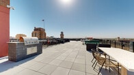 Located atop 705 Douglas Street in the heart of downtown Sioux City, Iowa, this rooftop terrace offers a compelling open-air experience elevated above the city skyline. The deck is finished with large-format neutral tile pavers that extend across the generous outdoor footprint, providing a clean, modern aesthetic underfoot.

Perimeter metal railings frame the entire rooftop, ensuring safety while preserving unobstructed 360-degree views of the surrounding cityscape. Standout landmarks visible from the terrace include a classic brick building flying the American flag and a neighboring clock tower, both characteristic of downtown Sioux City's historic architectural character. Expansive views of the broader city and surrounding landscape stretch to the horizon in every direction.

The terrace is equipped with a built-in outdoor kitchen station featuring a stainless steel grill and prep surfaces, making it well-suited for gatherings and events. Seating areas are distributed across the deck, including woven accent chairs, bench-style seating, and a substantial concrete pedestal table that serves as a central gathering point. Green modular seating adds a contemporary touch near the perimeter.

The brick exterior of the building's upper structure is visible along one edge of the rooftop, grounding the modern terrace in the building's traditional materiality. Abundant natural light and open sky above create a bright, airy atmosphere throughout.