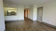 This single-floor space is designed with an open-concept layout that connects the main living area to the kitchen and beyond. The living area is finished with rich, dark hardwood-style flooring that runs throughout, offering a warm and cohesive aesthetic against the clean white walls.

The kitchen, visible from the main living space, features white cabinetry, a stainless steel stove, and a tile backsplash that adds a subtle decorative touch. An over-the-range microwave is also present, contributing to the kitchen's functional setup.

A hallway leads from the main living area toward additional rooms, with a warm-toned wood door visible at the end, suggesting access to a closet or private space. A six-panel door on the right side of the living area provides entry to another room within the unit.

Overhead ventilation grilles are integrated into the white ceiling, ensuring climate control throughout the space. The neutral color palette of white walls, gray baseboards, and dark flooring creates a clean, modern atmosphere that offers a versatile canvas for a variety of interior styles.