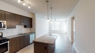 This single-floor unit showcases a contemporary design with a cohesive blend of dark espresso cabinetry, warm wood-tone flooring, and neutral walls throughout. The open-concept kitchen is a functional centerpiece, featuring dark-finish flat-front cabinetry extending to the ceiling, a white geometric tile backsplash, stainless steel appliances including a gas range, microwave, refrigerator, and dishwasher, and a spacious island with pendant lighting above. The kitchen flows seamlessly into a long, open living area illuminated by natural light from a large window and equipped with a ceiling fan. The living space benefits from light wood-tone laminate flooring and recessed lighting, creating a bright and airy atmosphere. A separate bedroom offers plush carpeting, a ceiling fan, and a window that invites natural light. The bathroom is well-appointed with large-format marble-look tile flooring, a deep soaking tub with a marble-surround shower enclosure, a wide vanity with dark cabinetry, a white countertop, and a large framed mirror. A dedicated in-unit laundry closet houses a stacked Whirlpool washer and dryer for added convenience. Track lighting in the kitchen and clean, white finishes throughout contribute to the unit's modern, move-in-ready appeal.