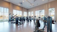 Located at 1749 Sea Pine Blvd in Myrtle Beach, SC, this fitness center is a thoughtfully designed, single-floor facility that offers an expansive and welcoming environment for exercise. The space is characterized by soaring ceilings with elegant tray detailing and ceiling fans that promote comfortable airflow throughout. Recessed lighting complements the abundant natural light that pours in through large, floor-to-ceiling windows lining multiple walls, offering views of the surrounding palm trees and the sunny South Carolina landscape. The open floor plan is generously proportioned, providing ample room between equipment for comfortable movement and accessibility. A full-length mirror along one wall enhances the sense of space and supports proper form during strength training. The durable, speckled flooring is well-suited for high-traffic fitness use. Equipment includes a comprehensive selection of strength training machines, treadmills, ellipticals, stationary bikes — including an Expresso interactive cycling bike — and cable machines. A dedicated storage and refreshment area with built-in shelving is also present. The overall ambiance is bright, clean, and professional, creating an inviting atmosphere suited for a wide range of fitness routines.