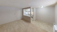 This single-floor unit offers a practical and well-organized layout designed for comfortable everyday living. The living area is generously proportioned, finished with neutral wall-to-wall carpeting and a textured ceiling that adds subtle character throughout. A wall-mounted air conditioning unit and baseboard heating ensure year-round climate control, while a large window invites natural light and provides views of the surrounding outdoor greenery.

A defining architectural feature is the open pass-through window connecting the living space to the kitchen, creating a sense of openness and continuity between the two areas. The kitchen is efficiently laid out with white cabinetry extending to the upper walls, a built-in range, refrigerator, and a sink set beneath additional cabinet storage. The tile flooring in the kitchen provides a clean contrast to the carpeted living zones.

The bedroom continues the neutral, light-filled aesthetic, featuring plush carpeting, a textured ceiling, a sliding closet with bifold doors, baseboard heating, and a window that looks out to a green outdoor area.

The bathroom is finished with classic white subway tile wrapping the tub surround, complemented by a small window that brings in natural light. The overall ambiance of the unit is clean, bright, and move-in ready, with a cohesive neutral palette throughout.