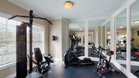 Located at 4121 Reserve Rd in Lexington, Kentucky, this dedicated fitness center is a well-appointed, single-floor exercise facility designed to support a comprehensive workout experience. The room is finished with warm beige walls, crisp white crown molding, and white-trimmed baseboards, lending a clean and polished aesthetic to the functional space.

A standout architectural feature is the expansive wall of framed floor-to-ceiling mirrors, which visually amplify the room and allow users to monitor their form during exercise. Natural light filters in through white slatted blinds on multiple windows, creating a bright and energizing environment. The durable dark rubber flooring provides both safety and comfort underfoot.

The fitness center is equipped with a well-rounded selection of Sole-brand cardio machines, including a treadmill, elliptical trainer, and spin bike, alongside a multi-function cable weight machine and additional free weights. A wall-mounted television is also present, offering entertainment during workouts. The layout is thoughtfully organized to allow comfortable movement between equipment.

Large interior windows connect the fitness room to an adjacent common area, which features warm hardwood flooring and elegant décor, reinforcing the upscale character of the broader facility. A posted fitness center rules sign near the entrance indicates this is a shared amenity space serving the surrounding community.