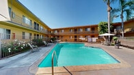 Located at 10800 Wright Road in South Gate, California, this residential complex is designed around a large, central outdoor swimming pool that serves as the focal point of the property. The complex features two-story building wings finished in warm yellow and orange tones, wrapping around the courtyard in a U-shaped configuration that creates a sense of community while providing direct pool views from multiple units.

Each unit is equipped with its own private balcony or ground-floor patio, offering residents convenient outdoor space with direct sightlines to the pool area. The balconies are fitted with metal railings and provide an elevated vantage point over the courtyard. Ground-floor units open onto a paved pool deck furnished with lounge chairs and a shaded seating area beneath a patio umbrella, ideal for relaxing in the Southern California sun.

The pool itself is generously sized with clear turquoise water, bordered by a fenced perimeter for safety. A staircase connects the two levels on at least one wing, offering easy access throughout the property. Mature palm trees and lush greenery punctuate the courtyard, adding a tropical ambiance that complements the region's warm climate. The overall design emphasizes outdoor living, natural light, and a resort-like atmosphere within a residential setting.