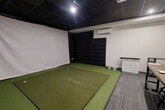 Located at 225 Sandwich St N in Amherstburg, Ontario, this purpose-built indoor golf simulator bay offers a well-equipped, self-contained environment for golf practice and entertainment. The space is designed around a central artificial turf hitting mat with multiple tee positions, positioned directly in front of a large white projection screen that spans the front wall — ready for simulator software integration via the ceiling-mounted projector overhead. Floor-to-ceiling black padded panels line the side and back walls, providing both impact protection and sound absorption while contributing to the room's sleek, modern aesthetic. The dark drop-tile ceiling with recessed and accent lighting creates a focused, immersive atmosphere. A ductless wall-mounted HVAC unit ensures comfortable year-round climate control. Along one side wall, a white cabinetry unit with a granite countertop provides convenient surface space, complemented by ball buckets nearby for easy access during sessions. The lounge and viewing area features a sturdy industrial-style communal table with adjustable stools, flanked by ball storage racks — making the space equally suited for group sessions or spectating. Patterned carpet tiles define the lounge zone, offering a clean visual contrast to the turf. A secondary door provides additional access to the space.