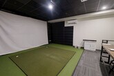 Located at 225 Sandwich St N in Amherstburg, Ontario, this purpose-built indoor golf simulator bay offers a well-equipped, self-contained environment for golf practice and entertainment. The space is designed around a central artificial turf hitting mat with multiple tee positions, positioned directly in front of a large white projection screen that spans the front wall — ready for simulator software integration via the ceiling-mounted projector overhead. Floor-to-ceiling black padded panels line the side and back walls, providing both impact protection and sound absorption while contributing to the room's sleek, modern aesthetic. The dark drop-tile ceiling with recessed and accent lighting creates a focused, immersive atmosphere. A ductless wall-mounted HVAC unit ensures comfortable year-round climate control. Along one side wall, a white cabinetry unit with a granite countertop provides convenient surface space, complemented by ball buckets nearby for easy access during sessions. The lounge and viewing area features a sturdy industrial-style communal table with adjustable stools, flanked by ball storage racks — making the space equally suited for group sessions or spectating. Patterned carpet tiles define the lounge zone, offering a clean visual contrast to the turf. A secondary door provides additional access to the space.