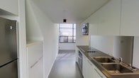 This single-floor space showcases a clean, contemporary design defined by a minimalist aesthetic throughout. The galley-style kitchen is a standout feature, offering a streamlined layout with crisp white cabinetry extending from floor to ceiling on both sides, maximizing storage while maintaining an uncluttered look. Stainless steel countertops lend a sleek, functional edge to the workspace, complemented by a double stainless steel sink and a built-in gas cooktop. A wall-mounted microwave and an oven are seamlessly integrated into the design for full culinary functionality. Light grey tile flooring runs the length of the kitchen, contributing to the space's cohesive, modern palette. A large window at the end of the kitchen floods the area with natural light, creating an open and airy atmosphere despite the narrow footprint. The window features horizontal blinds for adjustable light control. Recessed and flush-mount ceiling lighting ensures the space remains well-lit at all times. The overall ambiance is bright, efficient, and thoughtfully designed, reflecting a preference for clean lines and functional simplicity.