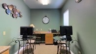 This single-floor space functions as a dedicated computer room, designed to accommodate shared or public use in a functional yet comfortable setting. The room is painted in a soft sage green, lending a calm, neutral atmosphere that promotes focus. Overhead, a flush ceiling light provides even illumination throughout the space, complemented by natural light filtering in through a window fitted with horizontal blinds on the right-hand wall.

The room is equipped with two glass-top bistro-style tables, each paired with ornate wrought-iron bar stools and topped with desktop computer monitors, making them ideal individual workstations. A warm wood-toned desk sits centered along the back wall, housing additional equipment including a printer, adding a practical administrative element to the space. A decorative oversized wall clock with Roman numerals serves as a focal point on the back wall, adding a touch of classic style to the otherwise utilitarian setting.

The carpeted flooring in a neutral beige tone adds warmth underfoot, while white baseboards provide a clean finish. The overall layout is efficient and organized, offering a welcoming environment for users needing computer access.