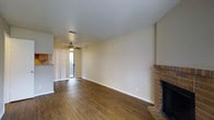 This two-story space offers a functional and comfortable layout with notable architectural details throughout. The main living area is a standout feature, showcasing rich, dark hardwood flooring that adds warmth and character to the room. A classic brick fireplace anchors one corner, serving as a focal point and providing both aesthetic charm and practical heating. Neutral, cream-colored walls create a clean and versatile backdrop, making the space easy to personalize.

Natural light filters into the living area through a sliding glass door fitted with vertical blinds, which opens to an outdoor area and enhances the sense of openness. A ceiling fan is positioned above, offering year-round comfort and air circulation. The adjacent kitchen area is partially visible, suggesting a convenient, open-concept flow between living and cooking spaces.

The two-story configuration provides additional square footage and potential for separated living and sleeping quarters. Overall, the space combines practical features with inviting design elements, including the warmth of the brick fireplace and hardwood flooring, creating a welcoming and livable environment.