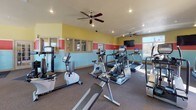 This fitness center is a comprehensively outfitted exercise facility designed to support a full range of workout routines. The space features a distinctive tri-color wall design — with bands of yellow, red, and teal — that gives the interior an energetic and motivating ambiance. Recessed lighting throughout and strategically placed ceiling fans ensure the space remains well-lit and comfortable during workouts.

The cardio zone is equipped with multiple treadmills, elliptical trainers, upright and recumbent stationary bikes, and additional aerobic machines. Wall-mounted flat-screen televisions are positioned for easy viewing from the cardio equipment. A full-length mirror spans one wall, visually expanding the space and allowing users to monitor their form.

The strength training area includes a variety of Cybex and other cable and selectorized weight machines, accommodating exercises targeting multiple muscle groups. A dedicated free weights section features a rack of dumbbells and flat benches, alongside stability balls and additional accessories.

Large windows along the perimeter fill the space with natural light and offer outdoor views, creating an open and inviting atmosphere. Durable rubber flooring throughout provides safety and comfort underfoot. The well-organized single-floor layout ensures smooth traffic flow between the cardio and strength zones, making this fitness center a functional and appealing facility.