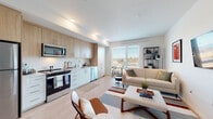 This well-designed single-floor residence offers a thoughtful layout that seamlessly connects its key living spaces. The entry hallway, finished with light wood flooring and crisp white walls, leads directly into the heart of the home. The open-concept kitchen and living area forms the centerpiece of the unit, where natural light pours in through large windows offering views of the surrounding neighborhood. The kitchen is outfitted with light wood cabinetry, white lower cabinets, a classic subway tile backsplash, and a full suite of stainless steel appliances including a range, microwave, dishwasher, and refrigerator. A cozy dining nook positioned beside a large window provides a bright spot for everyday meals. The living area offers generous space for seating and entertainment, anchored by light hardwood flooring and clean white walls throughout. The bedroom is a calm and private retreat, featuring soft, ambient lighting and a welcoming atmosphere. The bathroom is appointed with a wide white vanity, an undermount sink, a large LED-backlit mirror, and ample cabinet storage. A mini-split system provides climate control throughout. The overall design is modern and cohesive, with a neutral palette that promotes a bright, airy ambiance.